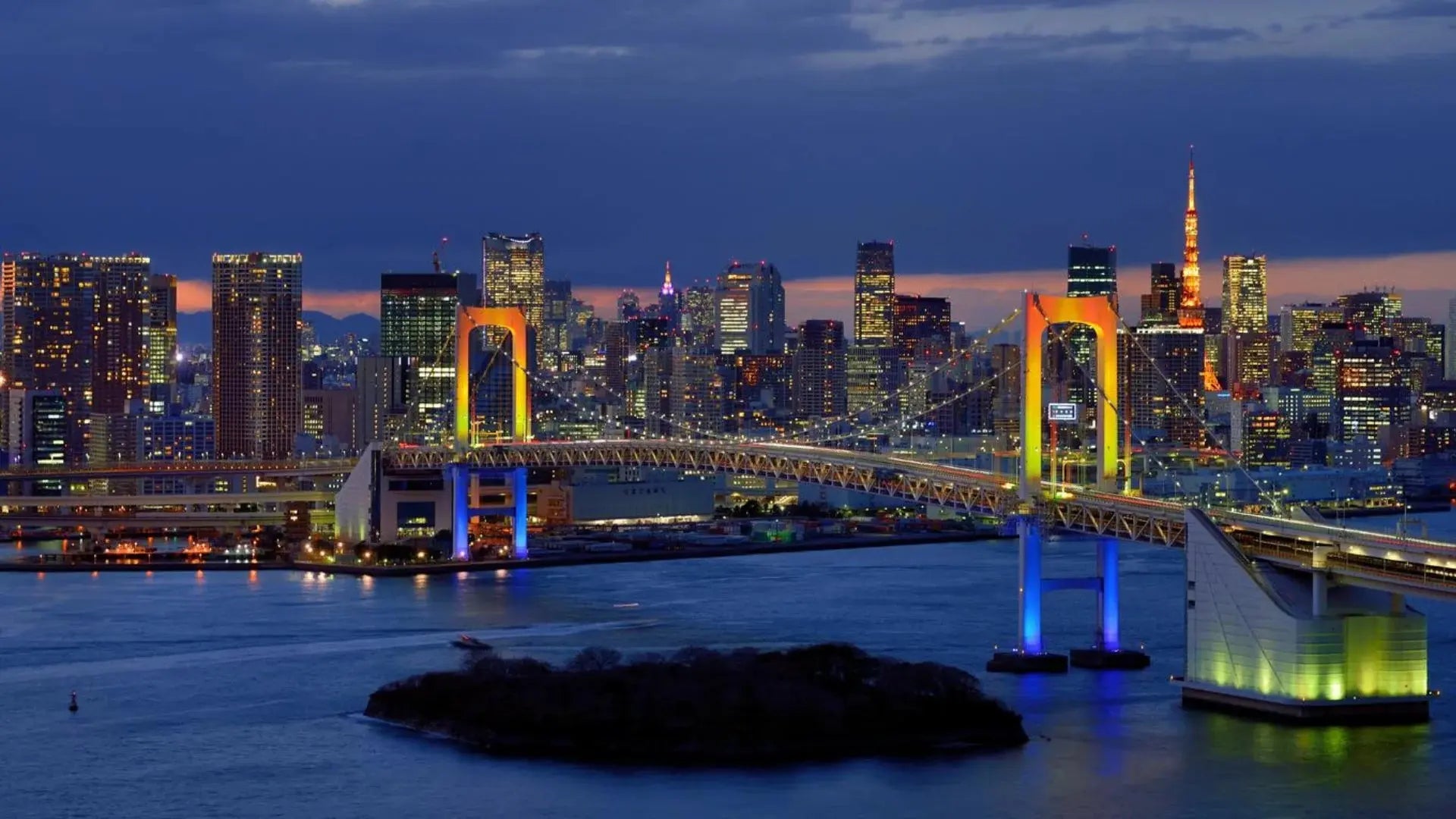 View of the iconic Rainbow Bridge in Odaiba, Tokyo, illuminated at night with vibrant colors. The bridge spans across Tokyo Bay, offering stunning views of the city skyline and waterfront